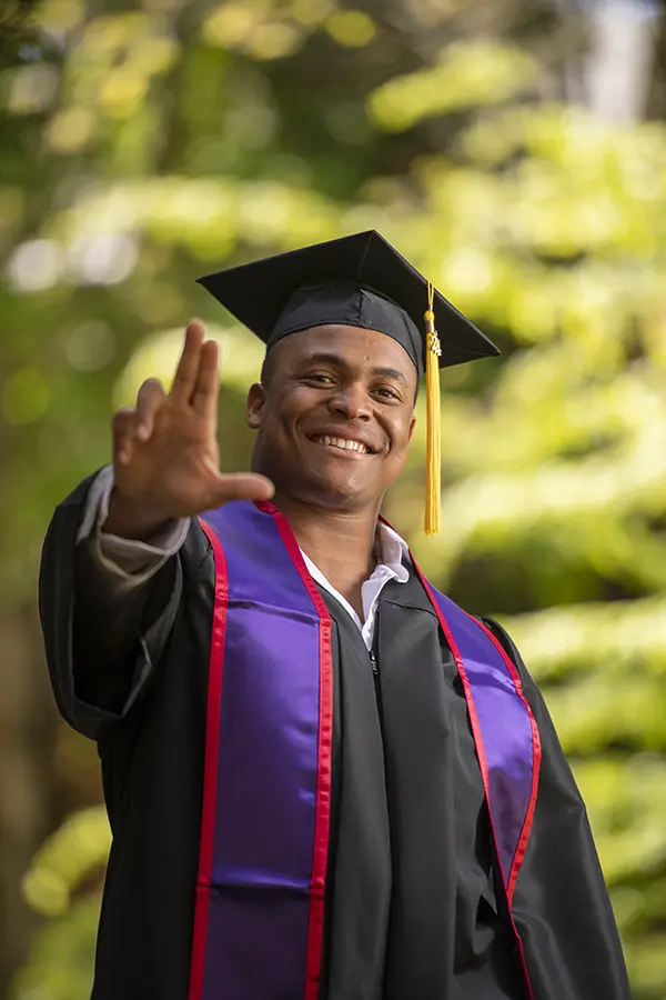 Divenson dressed in his graduation attire and making the Linfield L with his hand.